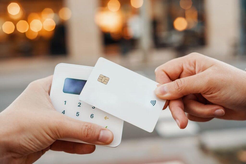 Foto close de mãos segurando maquininha e cartão por aproximação.