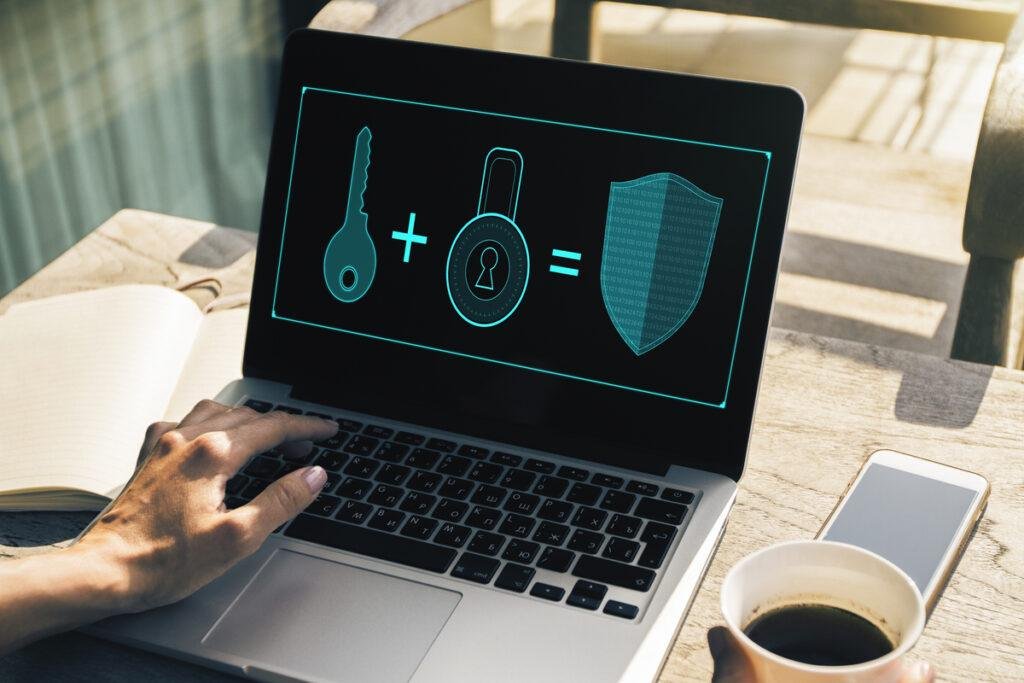 Foto de um computador sobre a mesa. Na tela há uma ilustração de chave + fechadura = escudo de segurança, representando a autenticação de dois fatores