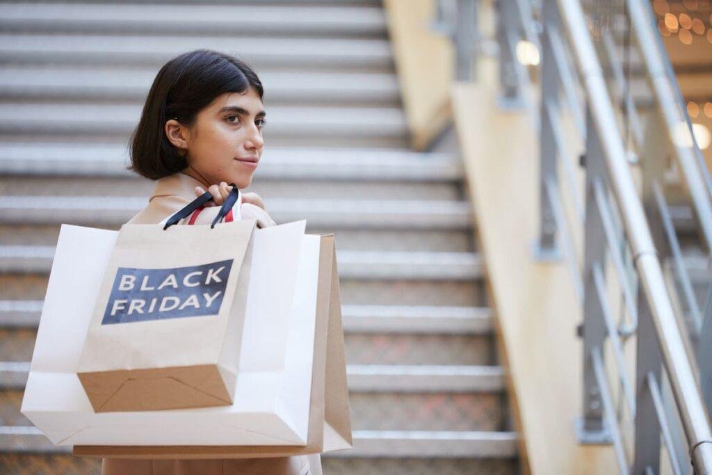 Foto de uma muher com sacolas de compras, uma delas escrito Black Friday, subindo uma escada de shopping