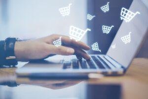 Close de uma mão acessando um notebook, carrinhos de compra flutuando sobre ele