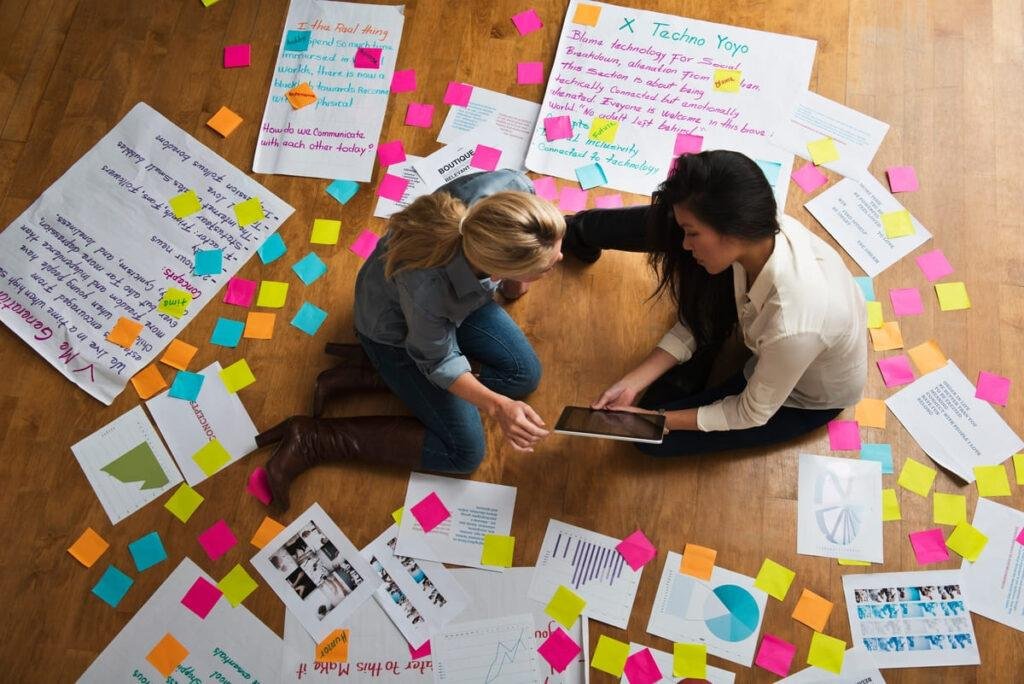Duas mulheres sentadas no chão, com grandes cartazes e post its espalhados