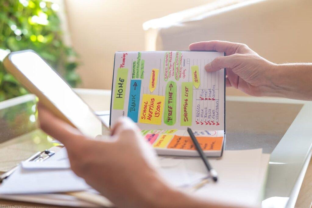 Foto de máos segurando uma agenda com post its coloridos representando prompts para organizar sua rotina